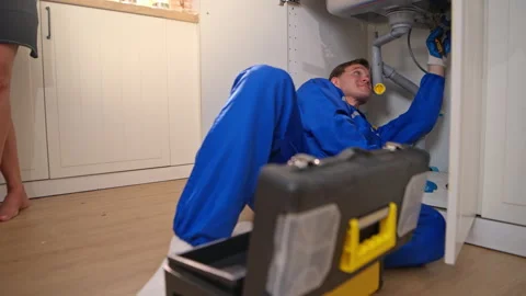Professional plumber fixing pipes under kitchen sink with toolbox nearby, Stock Footage 340183637
