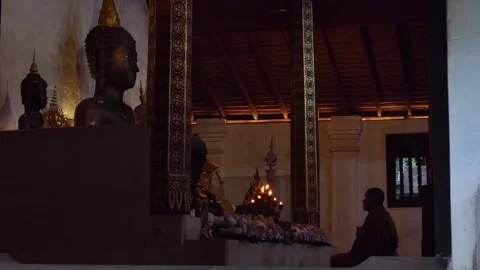 Profile view of monk praying in lotus position at altar with Buddha statues  Stock Footage 300882572