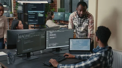 Programer sitting at desk with multiple screens running code talking with Stock Footage 168249054