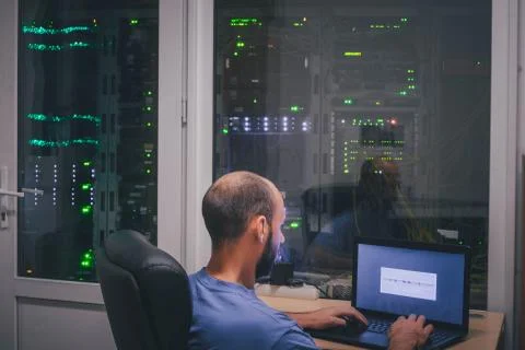 A programmer with a laptop sits in front of the glass wall of the server room Stock Photos