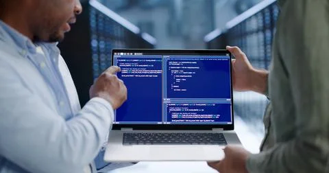 Programmer Reviews Secure Code On Laptop Within Busy Server Room. Stock Photos