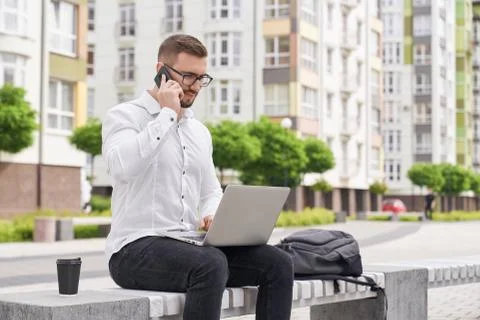 Programmer sitting on bench with laptop talking by phone. Stock Photos