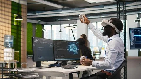 Programmer wearing VR headset at office desk developing programs using AI Stock Photos