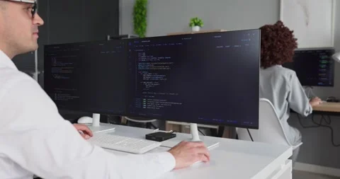 Programmer writing code on dual monitors in modern office with colleague nearby Stock-Footage 342024178