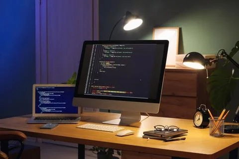 Programmer's workplace with computer, laptop and glowing lamps in dark office 스톡 사진