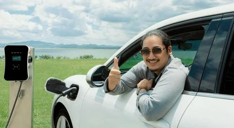 Progressive man connect power cable to his eco-friendly EV car on green field. Fotos Stock