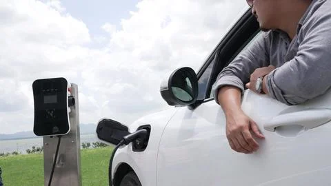 Progressive man connect power cable to his eco-friendly EV car on green field. Fotos Stock