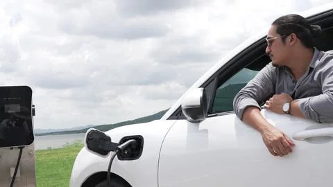 Progressive man connect power cable to his eco-friendly EV car on green field. Fotos Stock