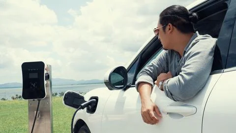 Progressive man connect power cable to his eco-friendly EV car on green field. Stock Photos