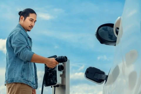Progressive man with plugged-in EV car with charging point, cloudscape backdrop. Stock Photos