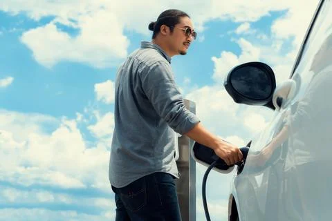 Progressive man with plugged-in EV car with charging point, cloudscape backdrop. Stock Photos