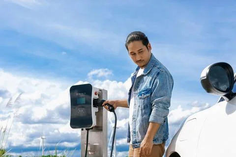 Progressive man with plugged-in EV car with charging point, cloudscape backdrop. Stock Photos