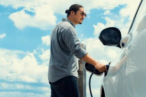 Progressive man with plugged-in EV car with charging point, cloudscape backdrop. Stock Photos