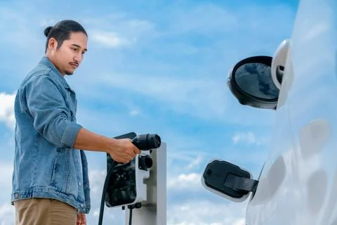 Progressive man with plugged-in EV car with charging point, cloudscape backdrop. Stock Photos