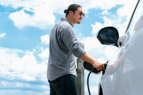 Progressive man with plugged-in EV car with charging point, cloudscape backdrop. Stock Photos