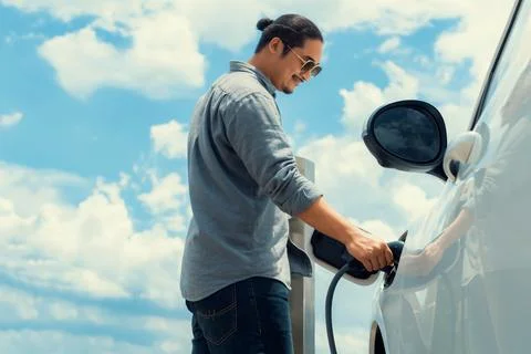 Progressive man with plugged-in EV car with charging point, cloudscape backdrop. Stock Photos