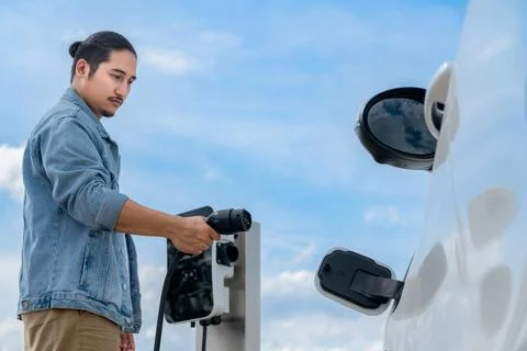 Progressive man with plugged-in EV car with charging point, cloudscape backdrop. Stock Photos