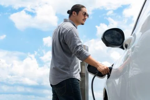 Progressive man with plugged-in EV car with charging point, cloudscape backdrop. Stock Photos