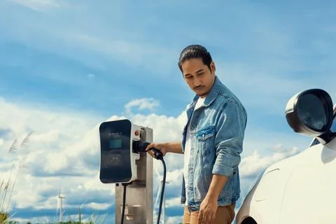 Progressive man with plugged-in EV car with charging point, cloudscape backdrop. Stock Photos