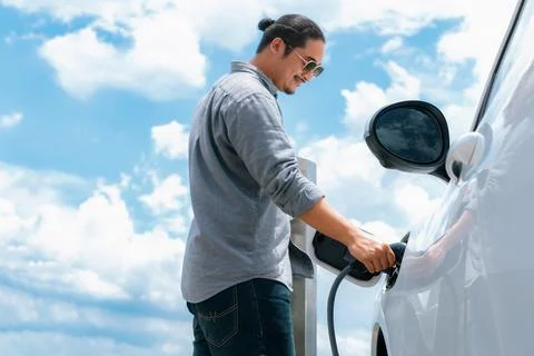 Progressive man with plugged-in EV car with charging point, cloudscape backdrop. Stock Photos