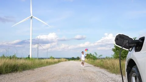 Progressive young boy playing windmill toy next to EV car at wind turbine farm. Stock Photos