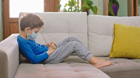 With a protective mask small boy with a smartphone sitting on the sofa and Stock Footage 127711850