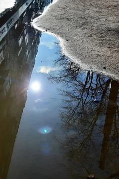 Puddle on the asphalt with the reflection of the blue sky and the bright sun. Stock Photos