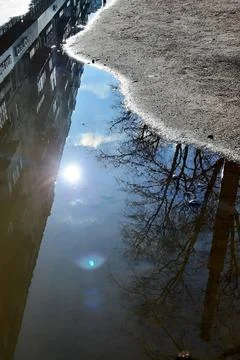 Puddle on the asphalt with the reflection of the blue sky and the bright sun. Stock Photos