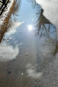 Puddle on the asphalt with the reflection of the blue sky and the bright sun. Stock Photos