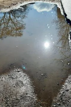 Puddle on the ground with a reflection of the blue sky and the bright sun. Stock Photos