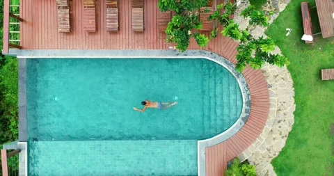 Pull-back aerial shot of man swimming in hotel's fancy pool on cloudy day. Stock Footage 278794385