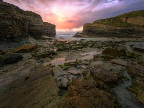Purple colors in the sunset clouds between the cliffs of a rocky beach Stock Photos