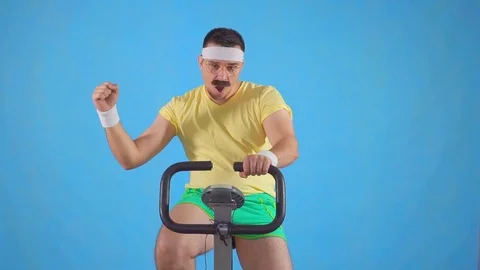 Purposeful athlete 80's with a mustache on a exercise bike on a blue background Stock Footage 107687286
