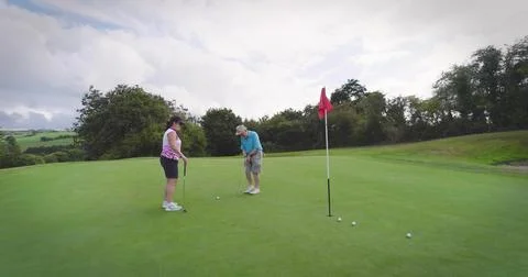 Putting man in light blue polo, woman in pink top holding putter on green, red Stock Photos