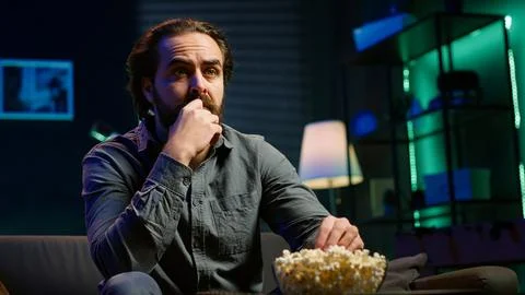 Puzzled man eating popcorn while watching confusing movie on TV Fotos de archivo