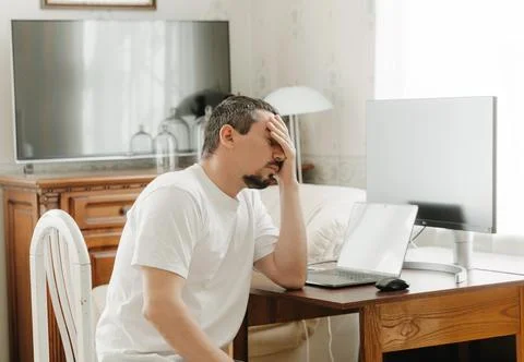 Puzzled man at a table with a computer with a hand near his face Stockfoto's