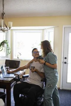 Quadriplegic man using laptop talking to medic girlfriend in scrubs Stock Photos