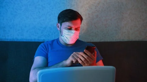 A quarantined masked man compares data from a phone and laptop. Video stock 129751143