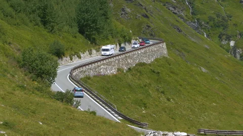 Queue of cars and white camper drive along alpine road bordered by green slopes Stock Footage 308336415