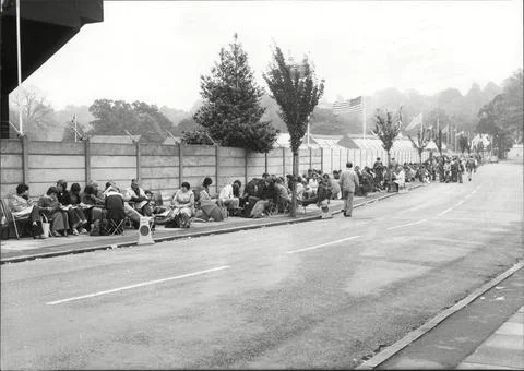 A Queue Of Tennis Fans Sit On The Ground As They Wait For The 1977 Wimbledon Ten Foto stock