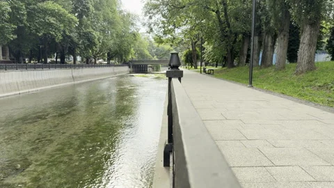 Quiet river flows past a paved embankment promenade lined with green trees Stock Footage 336938240