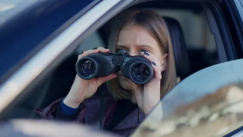 Rack focus from binoculars to serious focused young woman sitting in car spying Stock Footage 186289389
