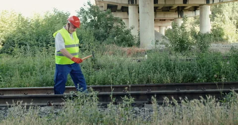 The railroader in uniform checks rails serviceability with hammer in summer day Stock Footage 135570754