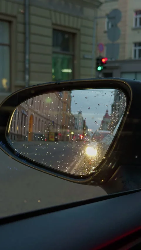 Rain covered car side mirror reflecting glowing city street traffic at dusk with Stock Footage 345129542