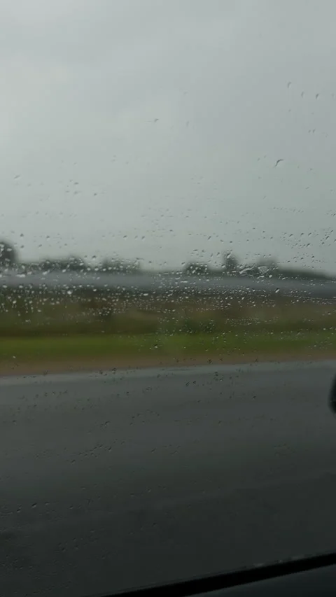 Rain droplets on car window with blurred roadside landscape under overcast sky Stock Footage 345763393