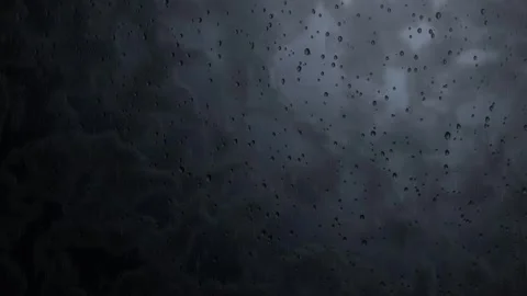 Rain droplets slide down a dark window pane against stormy clouds in a seamless Stock Footage 345375724