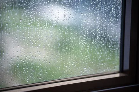 Rain droplets slide down windowpane, sunlight glimmers faintly outside. Ove.. Stock Photos