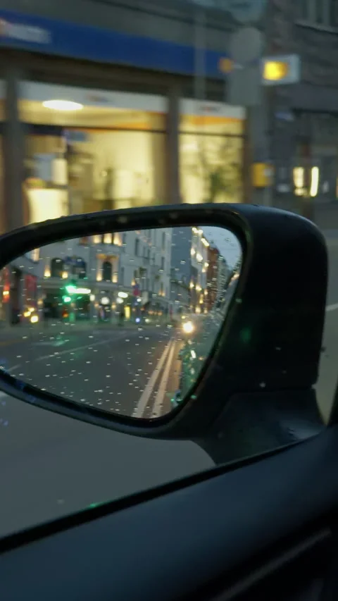 Rain-speckled side mirror reflecting blurred city street lights, wet buildings Stock Footage 344995773