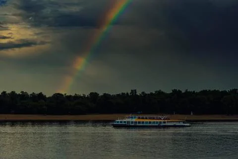 A rainbow from a dark cloud over a river and a motor ship. 스톡 사진
