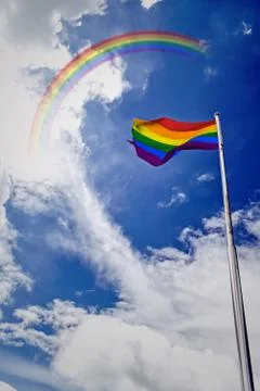 Rainbow flag waving in the wind under blue sky with rainbow. Stock Photos